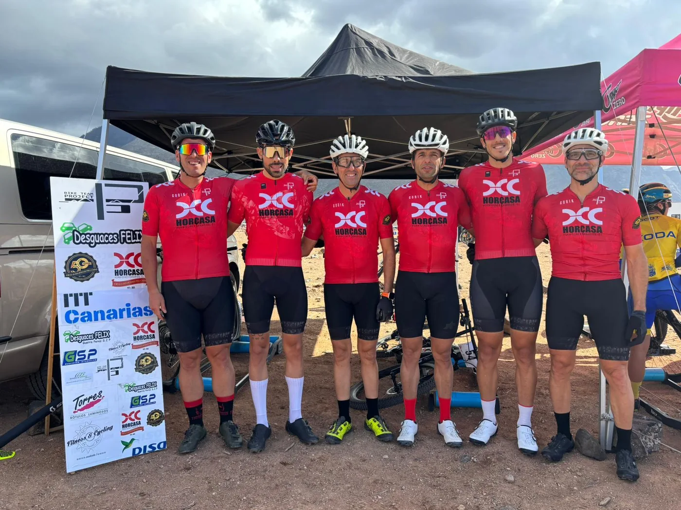 Seis ciclistas posan ante una carpa junto a un cartel de patrocinadores que incluye GSP